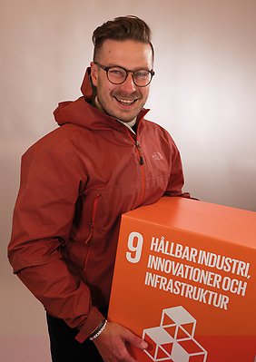 Max Östlund i en röd jacka håller i en röd låda med texten "Hållbar industri, innovationer och infrastruktur".
